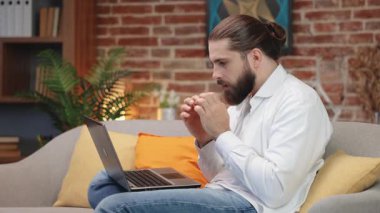 Hüsrana uğramış, uzun saçlı, çöreğe bağlanmış, dizüstü bilgisayarla koltukta oturan, sorun yaşayan bir adam. Evde uzaktan hareket halinde çalışırken hata yapan üzgün, şaşkın adamın yan görüntüsü. Duyguların kavramı.