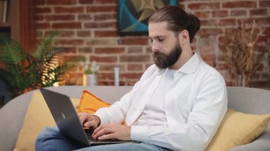 Evde dizüstü bilgisayarla çalışırken kameraya bakan çekici beyaz adam. Orta yaşlı yakışıklı erkek müdürün portresi koltukta oturuyor ve taşınabilir bilgisayarda çalışıyor. Uzak çalışma kavramı.