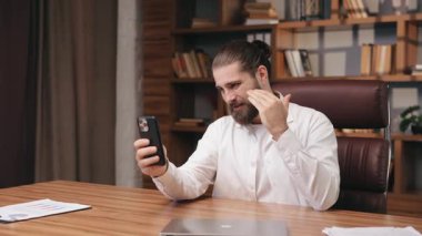 Resmi beyaz tişörtlü iddialı beyaz adam video toplantısı sırasında elinde akıllı telefon ile ofiste oturuyor. El kaldırıyor ve el kol hareketi yapıyor. Konsepti açıklamak ya da çalışanlara talimat vermek.