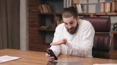 Ofisteki sinirli beyaz adam öne eğiliyor ve akıllı telefonu sıkıca tutuyor. Surat asmak ve hayal kırıklığıyla el kol hareketi yapmak. Yoğun görüntülü konuşma sırasında anlaşmazlık tartışmak veya ifade etmek.