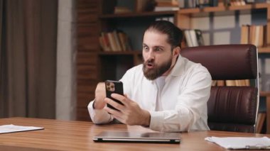 Beyaz tişörtlü kızgın sakallı bir erkek ahşap masada oturmuş, elinde akıllı telefon, video görüşmesi sırasında bağırıyor. Yumruğunu sıkarak ve kaşlarını çatarak masaya vurmak. Güçlü hayal kırıklığını ya da anlaşmazlığı ifade etmek.