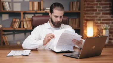 Saçları arkadan bağlanmış bir adam ahşap masada oturmuş dizüstü bilgisayarla çalışırken belgeleri dikkatle inceliyor. Ciddi bir erkek girişimcinin portresi evrak işlerine, çalışmaya ve harekete odaklanıyor. İş kavramı.