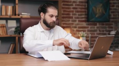 Orta yaşlı, odaklanmış bir adam ahşap masada oturuyor, dizüstü bilgisayarla çalışırken belgeleri inceliyor. Finans kontrolü yapan ciddi bir erkek girişimcinin portresi, rahat ofiste evrak işleri. İş, analitik kavram.