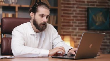 Gülümseyen sakallı serbest çalışan, günlük giyinen, içeride dizüstü bilgisayar üzerinde çalışırken masa başında oturan. Nişanlı erkek yöneticinin portresi, dizüstü bilgisayarda yazarken şaşırmış ve onaylanmış hissediyor. İş kavramı.