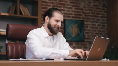 Sakallı iş adamı internette bilgisayar ekranına bakıyor. Beyaz tişörtlü, gülümseyen erkek yöneticinin portresi ofisteki meslektaşınla konuşmaya devam et. İş kavramı.