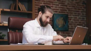 Odaklanmış beyaz gömlekli adam laptopta çalışıyor, pozitif sonuç aldığında sevinç hissediyor. Mutlu adamın yumruk kaldırması, dizüstü bilgisayara bakması, ahşap masada oturması. İş, başarı konsepti.