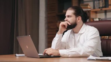 Uzun saçlı dalgın bir adam, masa başında oturmuş dizüstü bilgisayara bakarken derin düşüncelere dalmış. Sakallı iş adamının portresi dikkatle dizüstü bilgisayara odaklanıyor, çenesini uzatıyor. İş kavramı.
