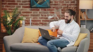 Kanepeye oturmuş, dizüstü bilgisayarı açık gülümseyen ve kollarını oynak bir şekilde hareket ettiren, günlük kıyafetli mutlu beyaz adam. Heyecan verici anı neşeli bir enerjiyle kutluyoruz. Eğlenceli bir şeye neşeli bir dansla karşılık vermek.