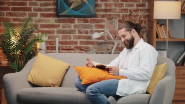 Beyaz gömlekli ve kot pantolonlu neşeli, uzun saçlı adam kanepede uzanıyor. Açık bir kitap tutuyor ve gülümsüyor. Sessizce gülerken öne doğru eğil. Keyifli, keyifli bir an yalnız, rahat bir oturma odasında.