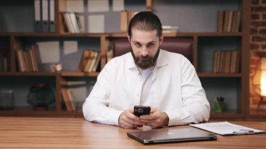 Beyaz gömlekli, kendine güvenen beyaz bir adam iki eliyle akıllı telefon tutuyor. Sakin bir ifadeyle doğrudan kameraya bakıyor. Göz teması kurarken dijital görev duraklatılıyor.