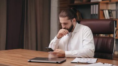 Ofisteki telefon ekranına bakarken ciddi bir orta yaşlı iş adamı düşüncesi. Düşünceli bir adamın portresi, akıllı telefon kullanırken rahat giyinen, çenesini ovuşturan. İş ve iş kavramı.