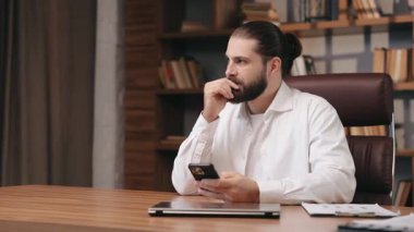 Masada otururken cep telefonu kullanarak düşünceli, sakallı girişimci düşüncesi. Beyaz gömlekli dalgın bir adamın portresi, akıllı telefona odaklanmış, çene ovuşturuyor, hareket halinde. İş kavramı.