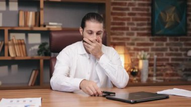 Beyaz gömlekli sıkıntılı beyaz bir adam sandalyeye yaslanıyor ve yüzünü iki eliyle kapatıyor. Akıllı telefon ekranı masaya yerleştirdi. Üzücü mesajlara tepki vermek ya da hayal kırıklığı yaratan haberler.