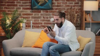 Akıllı telefonu sımsıkı tutarken öne doğru eğilen öfkeli beyaz adam. Şiddetli öfkeyle kaşlarını çatarak yumruğunu yastığa koyuyor. Sinir bozucu bir mesaj aldıktan sonra yatağa telefon atmak.