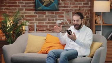 Beyaz gömlekli, koltukta oturan, akıllı telefona bakarken yumruğunu kaldıran heyecanlı beyaz adam. Ağız sevinçle bağırmak gibi açık. Çevrimiçi sohbet ya da bilgilendirme sırasında alınan inanılmaz haberlerden övgü.