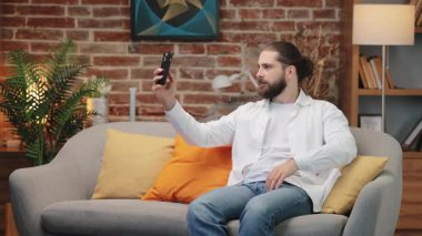 Beyaz gömlekli yakışıklı beyaz adam kanepeye oturmuş elinde akıllı bir telefonla barış işareti gösteriyor. Selfie çekerken kendine güvenerek gülümsüyor. Sıcak yaşam alanındaki neşeli anı yakalıyorum..