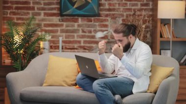 Beyaz gömlekli muzaffer beyaz adam koltukta bağdaş kurup heyecandan yumruklarını sıkıyor. Öne eğilip dizüstü bilgisayara gülümsüyor. Başarı kutlaması ya da internetten harika haberler almak.