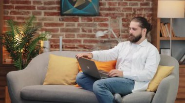 Kanepede dizüstü bilgisayarı kapalı oturan yorgun bir adam iki eliyle şakakları ovuyor. Uzun ekran süresinden sonra baş ağrısı belirtileri gösteriyor. Yorgunluk ya da zihinsel aşırı yüklenme ile mücadele.