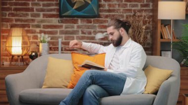 Uzun saçlı, düşünceli, sakallı bir erkek kanepeye yaslanıyor ve açık bir kitap tutuyor. Tamamen meşgul kalırken metin çalışıyor. Yaşam alanındaki anlamı yansıtmak ya da bilgiyi analiz etmek.