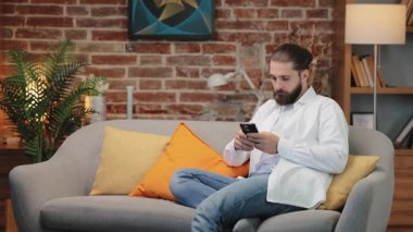 Odaklanmış beyaz erkek, düzgün sakallı, akıllı telefon tutarken kanepede bağdaş kurmuş oturuyor. Konsantre olurken dudaklara dokunmak. Önemli iletiler okunuyor veya mobil uygulamanın içeriği değerlendiriliyor.