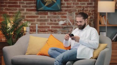 Samimi bir odada elinde akıllı telefon tutarak kanepede oturan uzun saçlı heyecanlı bir beyaz erkek. Gülümsüyor ve başarıyı kutluyor. Harika haberler okumak ya da anın tadını çıkarırken online ödül kazanmak.