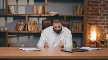 Beyaz gömlekli mutlu iş adamı, sıkı çalışma gününden sonra dans ediyor. Heyecanlı sakallı adamın portresi, rahat bir ofiste, ahşap masada otururken ofis sandalyesinde eğleniyor. Duygular, işyeri konsepti.