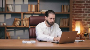 Laptop 'a bakarken dudaklarının yanında dinlenen düşünceli beyaz erkek yumruğu. Mesajı düşünmek ya da karmaşık bilgileri gözden geçirmek. Online görev sırasında sorun analiz ediliyor veya dikkatli tepki hazırlanıyor.