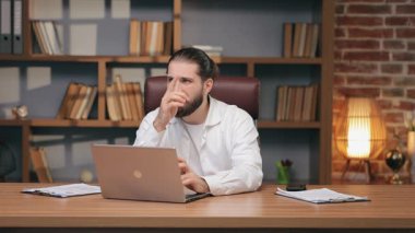 Ciddi bir erkek girişimci, sıcak bir ışıkla rahat ofisinde dizüstü bilgisayarla yazı yazıyor. Odaklanmış sakallı bir yönetici portresi, uzaktan çalışırken çenesini dinlendiriyor. İş ve iş kavramı.