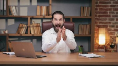 Heyecanlı beyaz erkek el çırpıyor ve masanın arkasında otururken gülümsüyor. Başarıyı kutlamak ya da sevinçle tepki vermek. Uzak toplantılarda başarı için tezahürat yapmak ya da heyecan verici haberler izlemek.