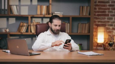 Saçları arkadan bağlanmış, ahşap masada oturan ve akıllı telefonları dinleyen odaklanmış beyaz bir adam. Kaynak taraması ya da güncellemeler kontrol ediliyor. İş görevlerini yok saymak ve online olarak zaman harcamak. Sıkıldım ya da kafam karıştı..