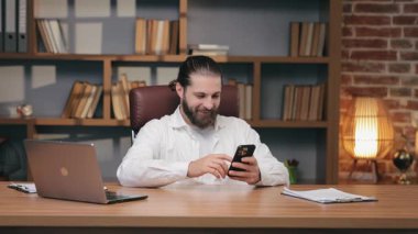 Sırtı bağlı, sırtı bağlı gülümseyen sakallı adam masada otururken akıllı telefonuna göz atıyor. Aydınlık anın tadını çıkarıyor ve sosyal medyayı kontrol ediyor. Komik içeriğe tepki vermek ya da iş molasında internette sohbet etmek.