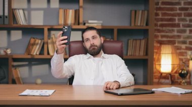 Beyaz tenli yakışıklı erkek akıllı telefonu yukarıda tutuyor ve parmaklarıyla barış işareti oluşturuyor. Samimi çalışma odasında selfie çekerken zorla gülümseme. Video çağrısı sırasında fotoğraf çekiliyor ya da paylaşılıyor.