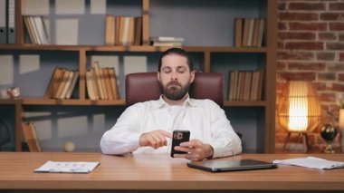 Beyaz tişörtlü sakallı sakin bir erkek akıllı telefon tutuyor ve kameraya doğru parmağını kaldırıyor. Modern iç mekanda masa başında otururken. İçerik onaylanıyor veya nötr yüzle ileti doğrulanıyor.