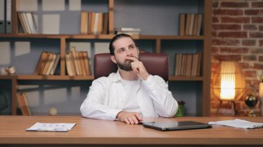 Saçları topuz şeklinde odaklanmış sakallı erkek. Kapalı dizüstü bilgisayarın yanında şık bir odada oturuyor. Yukarıya bakarken parmaklarını dudaklarına tutarak. İş sorununu göz önünde bulundurmak veya iş stratejisini yansıtmak.