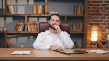 Beyaz gömlekli, sakallı ve sırtı bağlı düşünceli beyaz bir adam ofis masasında oturuyor. Dokunaklı çene ve ileriye bakıyor. Stratejiyi yansıtmak ya da önemli tepkiler hazırlamak.