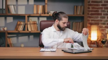 Kafkasyalı bir erkek dizüstü bilgisayarın yanında parmak izli çarşafları karıştırıyor. Yazılı sayfaları kontrol etmek ya da evrakları sıralamak. Ofis iş akışı sırasında birden fazla görev yürütülmesi veya belgelerin düzenlenmesi.