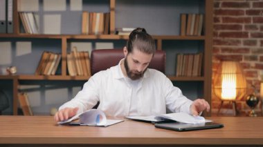 Masada otururken belgelere göz atan konsantre beyaz bir erkek. Belirli veriler aranıyor ya da finansal tablolara bakılıyor. Çalışma oturumunda raporlar hazırlanıyor veya rakamlar karşılaştırılıyor.