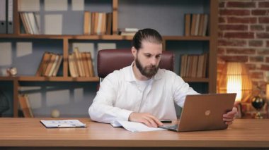 Ciddi bir beyaz adam masa başında otururken kağıtları kaldırıyor ve dizüstü bilgisayarda yazıyor. Çalışma oturumu başlatılıyor ya da sisteme giriş yapılıyor. Görevlerin tamamlanması veya dijital belgelerin etkin bir şekilde yönetilmesi için hazırlanılıyor.