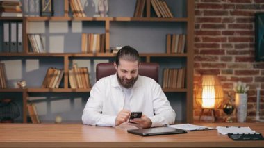 Beyaz tişörtlü beyaz erkek, akıllı telefon ekranında parmaklarını şaklatırken gülümsüyor. Rahatlamış mesajlaşma ya da mesajlar aracılığıyla gezinme. Sıcak ofisteki düzenli masanın tadını çıkarıyorum..