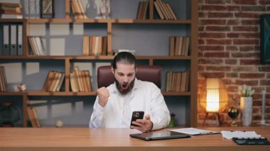 Akıllı telefona sevinçle bağırırken sıkılmış yumruğunu kaldıran heyecanlı beyaz adam. Başarıyı kutlamak ya da iş günü harika haberler almak. Modern ofis alanında zaferi ifade etmek.