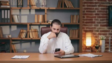 Akıllı telefona çatık kaşlı bir şekilde bakarken kafası kaşınan bir erkek. Organize ofis ortamında oturup mesajı analiz etmek ya da detayları hatırlamaya çalışmak. Bilgi ya da konu işleniyor.