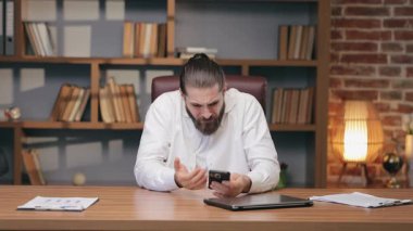 Beyaz gömlekli beyaz bir adam öne eğiliyor ve akıllı telefona somurtuyor. Üzücü haberleri okuduktan sonra inançsızlıkla el sıkışıyorsun. Masada tek başına otururken duygusal tepki veriyorsun..