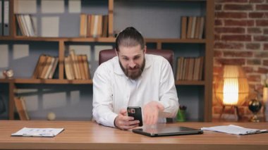 Şaşırmış sakallı erkek akıllı telefon tutarken şaşkınlık içinde el kaldırıyor. Masada oturup belirsiz bir mesaja ya da beklenmedik bir sonuca tepki vermek. Öfkeni ifade et ve açıklama iste..