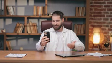 Beyaz gömlekli, gururlu, beyaz bir adam telefonu tutarken omzunun üstünden işaret ediyor. Video görüşmeleri sırasında yeni şık ofislerle caka satıyorsun. Sunulan arkaplan veya yakın zamanda elde edilen başarıları uzaktan paylaşma.
