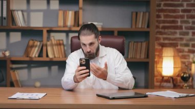 Şaşırmış sakallı adam başparmağını kaldırıyor ve kaşlarını kaldırmış akıllı telefona bakıyor. Çağrı sırasında mesaja tepki verirken masada oturmak. İş arkadaşı ya da takım arkadaşından beklenmedik haberler geliyor..