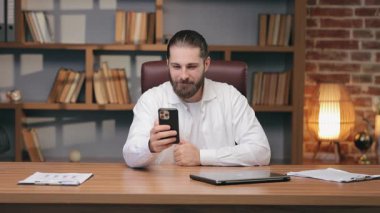 Beyaz tişörtlü neşeli sakallı erkek akıllı telefon tutuyor ve konuşurken gülüyor. Arkadaş ya da meslektaşımla gevşek bir görüntülü görüşme yapıyorum. Masada neşeli sohbetin tadını çıkarıyorum..