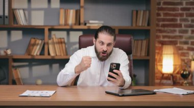 Masaya yumruk atarken akıllı telefona dik dik bakan asabi bir beyaz adam. Yoğun görüntülü konuşma ya da ateşli tartışma sırasında duygusal tepki vermek. Vücut dili ile güçlü anlaşmazlıkları ifade etmek.