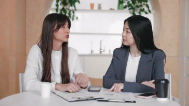 İki profesyonel arasında ortak masada iş meselelerini tartışan ciddi bir konuşma. Bir kadın belgelere doğru hareket ederken diğeri kollarını kavuşturarak dikkatle dinliyor..