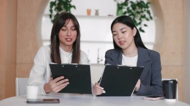 Takim arkadaslari masadaki notlari incelerken gündelik bir ses tonuyla sohbet ediyorlar. Belgeler, kahve ve mobil cihazlarla çevrili geri bildirim veya proje noktalarının netleştirilmesi.