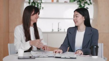 Gülümseyen kadınlar anlaşma ya da karşılıklı saygı jesti olarak el sıkışmak için masaya uzanırlar. Anlaşma yapmak ya da yeni ortaklığı kutlamak. Belgeler ve kahve fincanlarının yanında otururken..